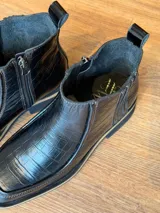 Botas cortas de cuero negro con textura de reptil, diseño de punta cuadrada y suela con detalle de franja en color beige.