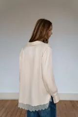 Camisa de satén color crema con cuello solapa, manga larga y cierre frontal con botones ocultos. Presenta un detalle de encaje en el bajo asimétrico.