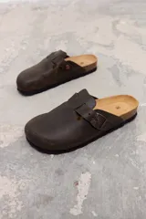 Zueco de cuero color marrón oscuro, con diseño de punta redonda, ajuste mediante hebilla metálica lateral y plantilla de corcho con suela de goma negra.