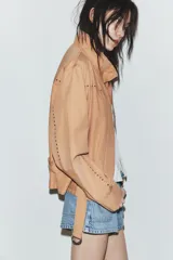 Chaqueta color tostado con cuello subido, manga larga y detalle de tachas metálicas en contraste. Incluye cinturón del mismo tejido con hebilla metálica y cierre frontal con cremallera y botones a presión ocultos.