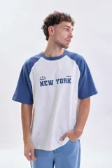 Remera de corte holgado con mangas ranglan en color azul y cuerpo en color blanco. Presenta un estampado frontal con la inscripción 'New York' y detalles gráficos.