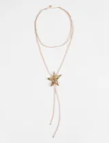 Collar de cordón doble color beige con dije de estrella metálica texturizada en tono dorado, que se ajusta mediante un nudo corredizo.