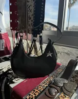 Cartera de hombro negra, con forma curva y cierre superior. Presenta un dije metálico plateado con forma de estrella en el frente. Incluye correa ajustable con ganchos metálicos.