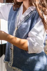 Chaleco tejido de punto color azul con destellos de lurex, escote en V y cierre frontal con botones. Presenta detalles de bordado artesanal en lana blanca en los bordes y terminaciones en crochet.