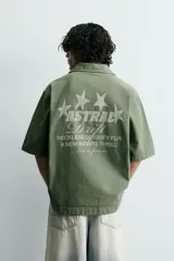 Camisa de corte relajado color verde oliva, con cuello solapa, manga corta y cierre frontal de botones. Presenta un estampado con el texto 'ASTRAL Drift' en el frente.
