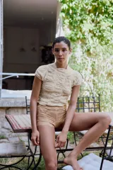 Top de punto con mezcla de viscosa, color beige, con cuello subido y manga corta. Presenta un diseño con frunces verticales y detalles bordados a tono, con terminación de ruedo ondulado.