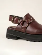 Zueco de cuero vacuno color bordó, con diseño de punta redondeada, detalle de tira con tachas metálicas y herrajes en tono oro viejo. Cuenta con un taco cuadrado de 4,5 cm y suela de cuero.