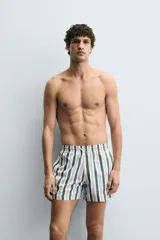 Bañador de hombre de tejido técnico con estampado de rayas verticales en verde oscuro, azul claro y blanco. Presenta cintura elástica ajustable con cordón blanco, bolsillos laterales y un bolsillo trasero de vivo.