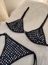 Conjunto de bikini negro con apliques de tachas cuadradas plateadas. El corpiño es de estilo triangular con una argolla central metálica y breteles regulables. La bombacha es de corte colaless con tiras laterales ajustables y argollas metálicas en los extremos.