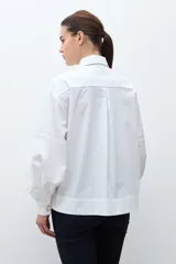 Camisa blanca de corte recto con cuello clásico, cierre frontal de botones y mangas largas con puños abullonados.