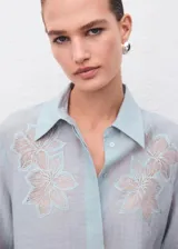 Blusa de tejido ligero color celeste con cuello camisero y cierre frontal de botones ocultos. Presenta mangas largas con puños anchos y detalles florales bordados en la parte superior delantera.