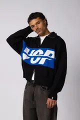 Campera tejida de punto con cierre frontal completo y cuello polo. Presenta un diseño con franja horizontal en color azul y letras en blanco sobre fondo negro, con puños y ruedo acanalados.