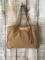 Cartera tipo tote de cuero con textura grabada tipo cocodrilo, color beige. Presenta un cierre frontal metálico en tono dorado y un detalle de borla decorativa colgante en el lateral. Cuenta con doble asa de hombro.