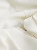 Blusa blanca de poliéster reciclado con cuello redondo, mangas cortas y cierre con cremallera en la espalda. Presenta una abertura en la parte delantera con barra metálica.