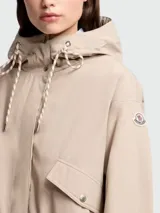Campera corta con capucha, color beige, de corte holgado y diseño minimalista. Presenta cierre frontal oculto, bolsillos laterales con solapa y botones a presión, mangas largas con puños ajustables y cordones decorativos en la capucha. Incluye el logo de la marca en la manga izquierda.