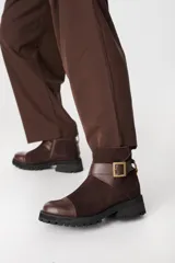 Bota corta de cuero color chocolate, con puntera reforzada, suela track de goma negra y detalle de tira con hebilla metálica en el tobillo.