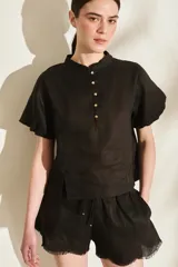 Blusa negra de lino con cuello mao, mangas cortas abullonadas y cierre frontal parcial con botones dorados. Presenta pequeños tajos laterales en el ruedo.
