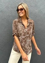 Camisa de corte recto y manga corta con cuello clásico y cierre frontal de botones. Presenta un estampado floral en color blanco sobre una base de color chocolate.