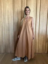 Vestido largo color camel, sin mangas, con escote en V y costuras verticales decorativas. Presenta bolsillos laterales y aberturas en los costados de la falda.