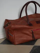 Bolso de viaje tipo weekender de cuero color tostado con asas y detalles en marrón oscuro, herrajes metálicos y bolsillo frontal con cierre.