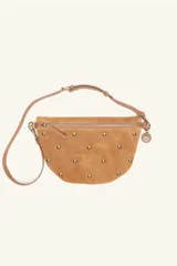 Bandolera de gamuza color tostado con diseño semicircular, decorada con tachas metálicas doradas distribuidas en el frente. Cuenta con cierre superior, correa ajustable y un dije colgante metálico.