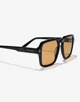 Lentes de sol Hawkers modelo Lash Eco en colaboración con Smiley. Presentan un marco cuadrado de color negro con acabado brillante y cristales en tono ámbar. Incluyen detalles metálicos en las esquinas y el logo de Smiley grabado en las patillas.