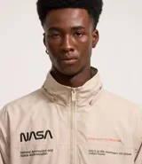 Campera rompevientos color beige con capucha, cierre frontal y bolsillos laterales. Presenta estampado de la NASA en el pecho, parche circular en la manga derecha y bandera de Estados Unidos en la manga izquierda.