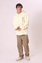 Buzo con capucha color crema, de corte holgado, con bolsillo frontal tipo canguro y pequeña estampa gráfica en el pecho.