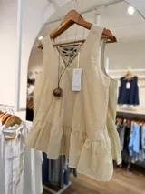 Blusa sin mangas color beige de silueta fluida, con escote en V y volado peplum en el ruedo. Presenta espalda con tiras cruzadas ajustables y viene acompañada de un collar con dije redondo de diseño rústico.