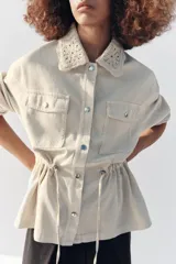 Chaqueta estilo safari color beige, con cuello de solapa tejido en crochet, manga larga, bolsillos delanteros con solapa y cintura ajustable con cordones. Cierre frontal con botones metálicos a presión.
