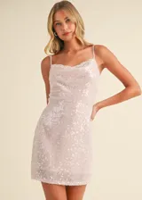 Vestido corto de tirantes finos, confeccionado en tela con lentejuelas brillantes en tono rosado claro. Presenta un escote tipo cowl con un delicado detalle de encaje en el borde superior y una silueta ajustada.