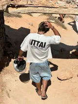 Remera marrón de corte holgado con estampado en la espalda que incluye la palabra "ITTAI" en blanco con líneas rojas y azules, la frase "AN IBIZA PRODUCE" y un texto más pequeño que dice "Ittai Stars Department Gallery - Ibiza Edition. If you're in Ibiza, 112 is not an option.".