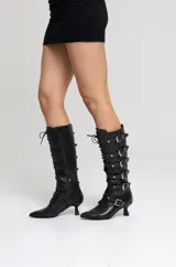 Botas altas de cuero negro con acabado brillante texturado, múltiples tiras con hebillas y tachas plateadas, cordones frontales, punta fina y taco bajo escultural.