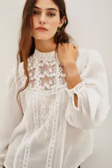 Camisa blanca de estilo romántico con cuello alto, confeccionada en algodón con detalles de encaje y bordados en el frente. Presenta mangas largas abullonadas con puños elásticos y terminaciones de encaje.