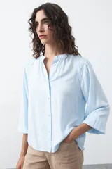 Camisa de color celeste claro con silueta relajada y mangas largas. Presenta un cuello redondo con pequeña abertura frontal y botones, además de un delicado trabajo de tablas en los hombros que aporta textura al diseño.