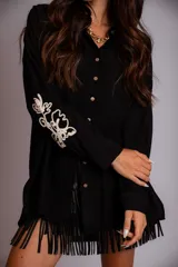 Camisa beige de corte amplio, con cuello clásico, cierre frontal con botones, mangas largas con puños abotonados y bordado floral en las mangas.