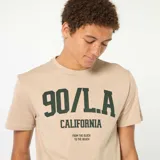 Camiseta de algodón color beige con cuello redondo y mangas cortas. Presenta un estampado frontal en color verde oscuro con la inscripción '90/L.A. CALIFORNIA' y el texto 'FROM THE BLOCK TO THE BEACH'.