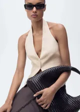 Chaleco sastrero de lino color beige, con cuello halter pronunciado y cierre frontal de cuatro botones. Presenta bolsillos ribeteados simulados en el frente y dobladillo en punta.