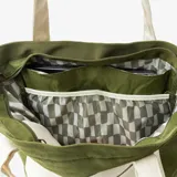 Bolso tote verde militar confeccionado en nylon resistente al agua, con un bolsillo frontal grande en color crudo que incluye cierre horizontal y una anilla metálica. Cuenta con asas largas de hombro en color crudo y una manija superior corta.