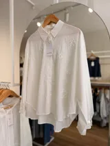 Camisa oversize blanca de manga larga, con cuello camisero, cierre frontal de botones y delicados bordados florales tono sobre tono.