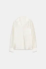 Camisa de cuello solapa y escote pico, manga larga, color blanco roto. Presenta un bolsillo plastrón en el delantero con bordado combinado a tono y bajo asimétrico.