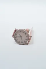 Reloj analógico con correa de cuero sintético color rosado pálido y caja a juego. La esfera es beige claro con números romanos en negro.