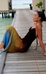 Vestido largo de seda o material similar, con tirantes finos y escote drapeado tipo cowl neck. Presenta un efecto degradado de color que va del negro/marrón oscuro en el busto, pasando por tonos naranjas y amarillos, hasta terminar en un azul vibrante en el ruedo.