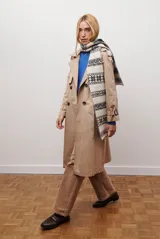 Trench coat color beige con cuello de solapa, cierre frontal abotonado, bolsillos laterales y mangas largas con cintas ajustables. Incluye capucha desmontable y forro interior rayado. La tela exterior cuenta con protección repelente a la lluvia y manchas.