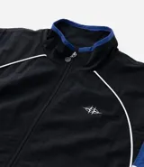 Campera deportiva de poliamida con diseño de bloques de color en negro, blanco y violeta. Presenta cuello alto, cierre frontal con cremallera, puños y ruedo elásticos, y forro interior de poliéster.