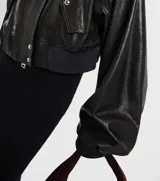 Campera bomber corta de cuero sintético negro, con cuello alto, cierre frontal y detalles de botones metálicos plateados en hombros y bolsillos frontales.
