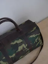 Bolso de viaje tipo duffel de lona negra con detalles en cuero tostado, asas de mano y correa ajustable para el hombro.
