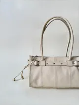 Cartera de cuero de tamaño medio con estructura rígida. Presenta una tira decorativa con tachas y hebillas metálicas en los extremos, además de lazos laterales. Cuenta con doble asa para llevar al hombro y cierre superior.