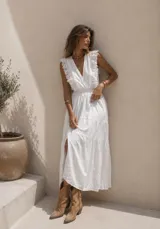 Vestido largo blanco de broderie, sin mangas, con escote en V profundo y volados en los hombros. Presenta cintura elastizada fruncida y falda larga con capas escalonadas.