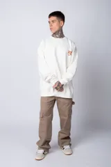 Buzo de tejido friza color crema, de corte oversize y cuello redondo. Presenta un pequeño bordado o parche en el pecho izquierdo con un diseño de cabeza de animal en tonos rojizos/terracota.